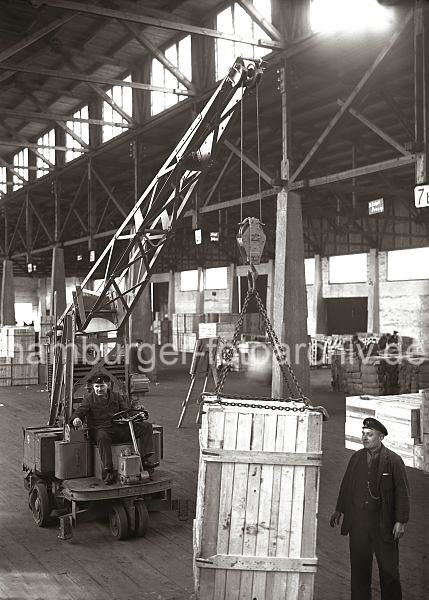 Transport einer Holzkiste mit einem Elektrokran; ca. 1932. Alte Fotografien von der Arbeit im Hamburger Hafen aus dem - Bildarchiv der HHLA. 638_58 Eine gro�e Holzkiste wird in einer der vielen Lagerhallen des Hamburger Hafens mit einem Laufkran transportiert. Vier Metallkrallen, die sich an einer Eisenkette des Kranhakens befinden, tragen die Kiste, die grob aus Holzbrettern zum Schutz des Transportguts zusammengezimmert ist. Der Fahrer des Elektrokrans hat eine Hand am Lenkrad, w�hrend die andere auf den Hebeln liegt, mit denen die Kiste am Haken bewegt wird. Ein weiterer Arbeiter dirigiert den Transport zu seinem Ziel.