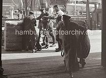 670_727 Ein Kaiarbeiter zieht seine Sackkarre mit einem schweren Ballen von der Rampe in den Lagerschuppen. Im Hintergrund sind drei Arbeiter dabei, einen der Ballen auf eine Sackkarre zu setzen.