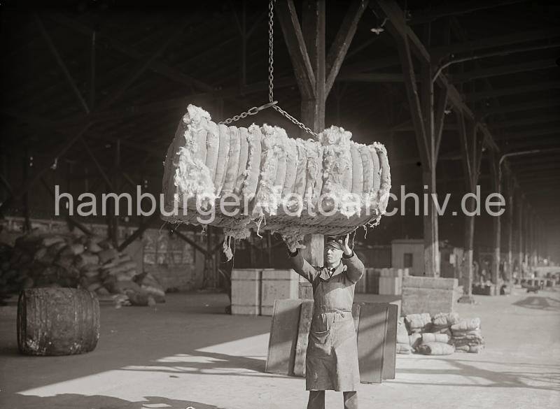 672_439 Ein Wollballen hngt im Ladegeschirr und wird von einem Hafenarbeiter am Kai angenommen. Im Hintergrund sind in dem offenen Schuppen Kisten und Scke gelagert. Hafenfotos  aus dem Bild-Archiv  der Hamburger Hafen und Logistik AG (HHLA) - Verladung eines Wollballens am Hafenkai - Hafenarbeiter mit Schrze und Dienstmtze; ca. 1934. 