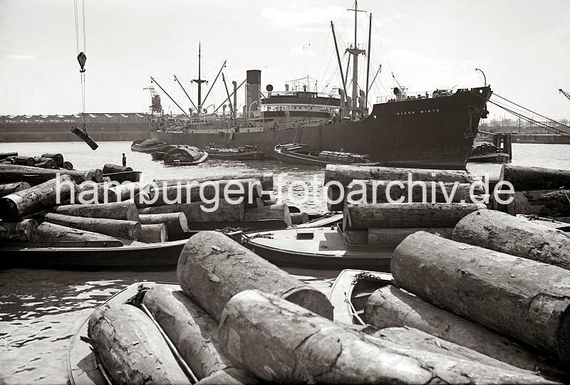 Historische Fotografien zur Hafengeschichte Hamburgs aus dem Archiv der HHLA. Tropische Baumstämme werden auf Schuten geladen; ca. 1938.  692_395a Die Stämme tropischer Bäum werden auf Schuten geladen - einer der Baumstämme hängt in einer Baumzange am Kranhacken und wird auf den Lastkahn herunter gelassen. Im Hintergrund liegt das Frachtschiff BARON MINTO an den Holzdalben; die Ladung wird mit schiffseigenen Ladebäumen auf auf Schuten gelöscht .