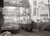 699_585a Mit Holzbohlen beladene Güterwaggons sind am Kai auf den Gleisen vor den Frachter geschoben worden - mit dem Kran werden die Bündel Holzbretter von den offenen Waggons gehoben und auf das Schiff transportiert.
