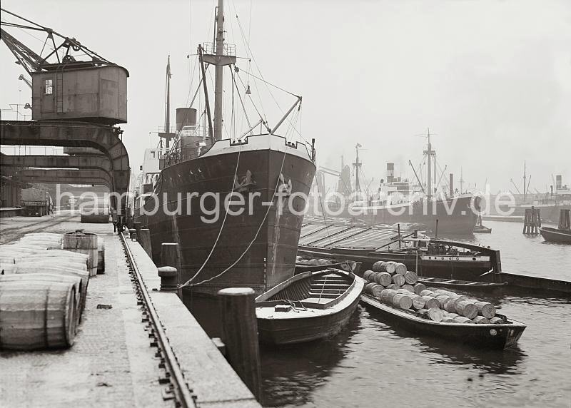 Historische Hafenbilder aus Hamburg - Bilddokumente aus dem Archiv der HHLA. Frachtschiff + Schuten / Binnenschiff am Hafenkai.  01_701 Eine Ladung von Holzfssern wird von Bord eines Frachters gelscht. Auf den lngsseits liegenden Schuten werden die Tonnen verladen - ein Elbkahn mit enormen Steuerruder liegt ebenfalls an der Wasserseite des Frachtschiffs. Im Hintergrund wird an den Dalben die Fracht eines Schiffs mit einem Getreideheber gelscht.