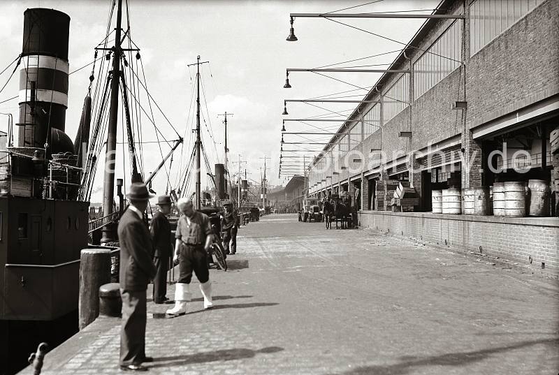 Bilder Altonas - Fischdampfer am Kais des Altonaer Fischereihafen, Pferdefuhrwerk / Zugmaschine + Anhnger; ca. 1937. AltonaMotive als Kunstdruck auf Leinwand + Poster.  804_B_287 Rechts die 1937 neu gebaute Fischauktionshalle am Kai des Altonaer Fischereihafens -  Tonnen und Holzkisten zum Transport der Fische stehen auf der Laderampe des Gebudes. Ein Pferdefuhrwerk wird beladen, dahinter ein Zugmaschine mit Anhnger, die von Kaiarbeitern mit Kisten beladen wird. Mehrere Fischdampfer mit hohem Schornstein liegen am Kai - einer der Fischer geht mit heruntergeklappten Fischer-Gummistiefeln ber die Kaianlage.