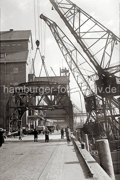 Abbau eines 40t Doppel-Lenker Wippkrans; 1938 814_B_358a Durch den Anschluss Altonas an Hamburg 1936 wurden auch die beiden Hfen Hamburg. und Altonas koordiniert. In diesem Zusammenhang wird der 40t Doppellenker Wippkran am Kai des Altonaer Hafens abgebau und am Rohft wieder aufgestellt. Zwei Schwimmkrne hieven das Unterge- stell der Krananlage an. Im Hintergrund stehen an der Laderampe des Getredesilos beladene Lastwagen und Gterwaggons.