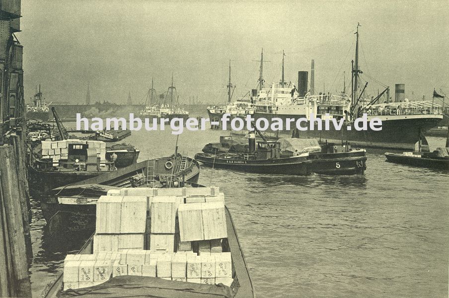 0953983 BIm Hansahafen werden Schuten beladen - hoch getrmt liegen Holzkisten in einem Kahn; ein anderes Transportschiff wird mit seiner Ladung von einem Schlepper zu seinem Bestimmungsort gebracht. Im Hintergrund Kirchtrme der Hansestadt Hamburg.