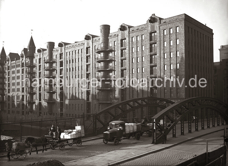 Alte Fotografien von der Hamburger Speischerstadt aus dem Fotoarchiv der HHLA, Gterverkehr auf der Poggenmhlenbrcke - Speicherblock W; ca. 1930. 01147755_166 ber die Poggenmhlenbrcke, die ber den Hollndischen Brooksfleet fhrt, werden Gter in die Speicherstadt transportiert. Im Vordergrund zieht ein Transportarbeiter eine  Schottsche Karre mit Paketen, dahinter sitzen zwei Kutscher auf dem Bock eines Pferdefuhrwerks, das Holzkisten geladen hat. Ein Lastkraftwagen mit Anhnger hat Holztonnen auf seiner Ladeflche und es folgt noch eine weitere Handkarre mit Waren.