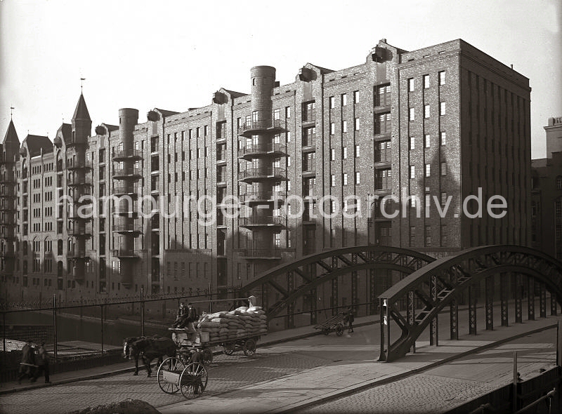 Alte Hamburg-Motive von der Speischerstadt aus dem Fotoarchiv der HHLA - Poggenmhlenbrcke, Pferd und Wagen + Speicherblock W; ca. 1930.  01147756_166a ber die Poggenmhlenbrcke, die ber das Hollndischen Brooksfleet / Wandrahmsfleet fhrt, transportiert ein Pferdfuhrwerk mit zwei Kutschern eine Ladung Scke; die Plane, die auf eine Stange ber die Ladung gelegt werden kann, ist zurck gezogen. Im Vordergrund ein Arbeiter mit seiner Karre zur Straenreinigung(?) mit Besen und Schaufel auf der Ladeflche.