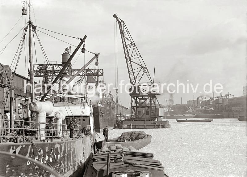 Historische Fotodokumente aus dem Archiv der HHLA Frachtschiffe im Sandtorhafen / Sandtorkai (?), Schwimmkran; - alte Hamburg Fotografien auf Leinwand oder Posterprints.  01180_533 Ein Schwimmkran mit einer Holzkiste am Haken liegt lngsseits eines Frachters im Sandtorhafen (?) - das Wasser des Hafenbeckens ist eisbedeckt. Im Vordergrund ist der Frachter DELPHIN, beladene Schuten liegen lngsseits.